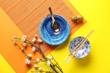 Bamboo mat with bowls, chopsticks and sakura branch on yellow background. Chinese table setting