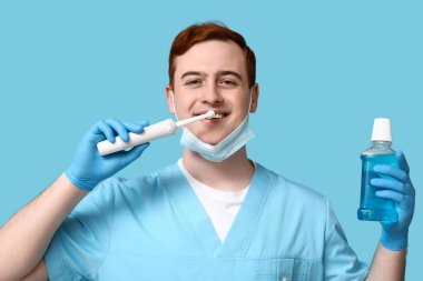 Male dentist with electric toothbrush and bottle of mouth rinse on blue background. World Dentist Day