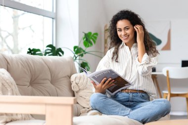 Young African-American woman reading magazine on sofa at home