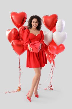 Young African-American woman with gift and balloons on light background. Valentine's Day celebration