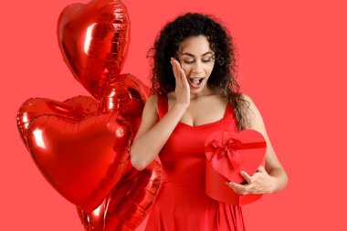 Surprised young African-American woman with gift and balloons on red background. Valentine's Day celebration