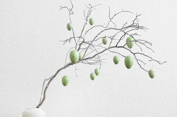 Tree branch decorated with Easter eggs against light wall, closeup
