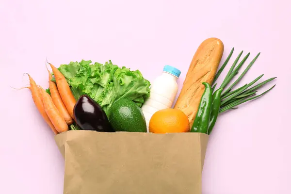 Paper bag with vegetables, milk and bread on pink background