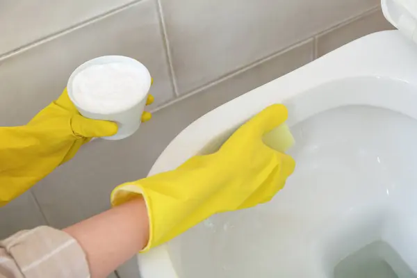 Woman in rubber gloves cleaning white toilet bowl with baking soda and sponge, closeup