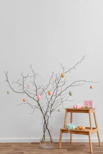 Tree branches in vase and stepladder with Easter eggs near white wall