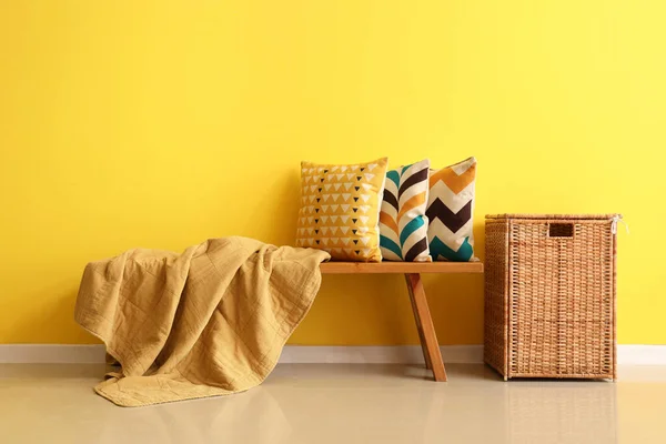 Wooden bench with cushions and wicker basket near yellow wall