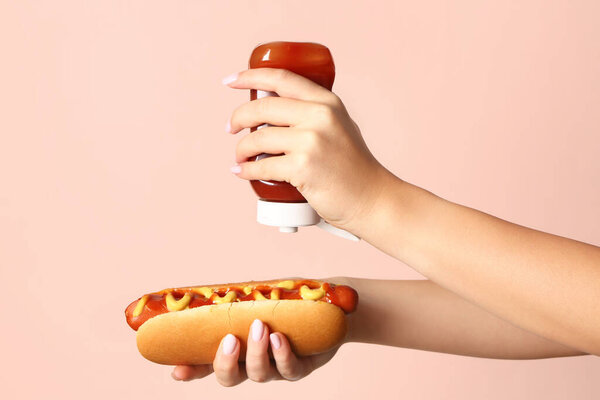 Woman adding ketchup onto tasty hot dog on pink background