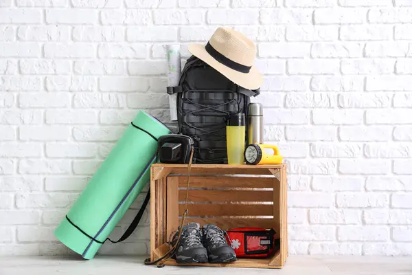 Set of camping equipment with backpack, flashlight, thermos and hat near white brick wall