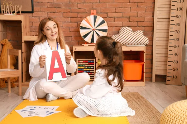 Mature speech therapist teaching little girl how to pronounce letter A in office