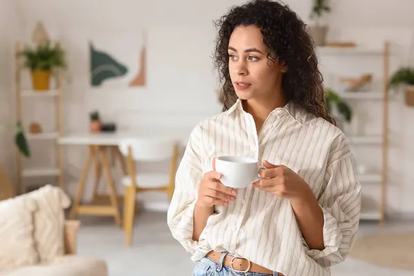Young African-American woman with cup of coffee at home