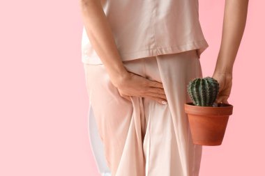 Young woman in pajamas with hemorrhoids and cactus on pink background, back view