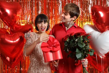 Young woman opening gift box from her boyfriend against tinsel on red background. Valentine's Day celebration