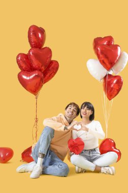 Young couple with gift box and balloons sitting on yellow background. Valentine's Day celebration