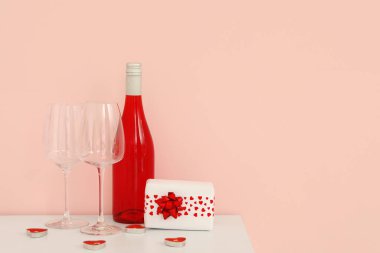 White cabinet with bottle of wine, glasses, gift box and heart-shaped candles near pink wall. Valentine's Day celebration