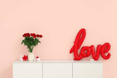 White cabinet with balloons in shape of word LOVE, gift boxes and bouquet of roses near pink wall. Valentine's Day celebration