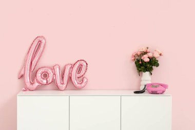 White cabinet with word LOVE made from balloons, bouquet of roses and vintage telephone near pink wall. Valentine's Day celebration