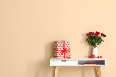 White desk with bouquet of roses, gift box and candles near beige wall. Valentine's Day celebration