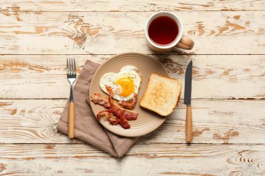 Plate with tasty bacon, cup of tea, toast, fried egg and cutlery on light wooden background