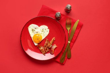 Plate with tasty bacon, heart made of fried egg and cutlery on red background