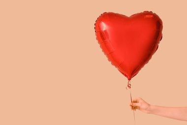 Female hand holding heart-shaped balloon on beige background. Valentine's Day celebration