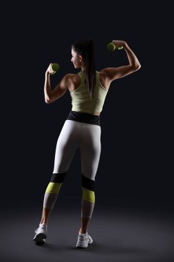 Sporty young woman exercising with dumbbells on black background, back view