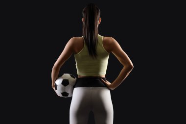 Sporty young woman with soccer ball on black background, back view