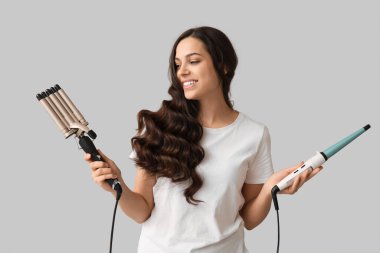 Beautiful young woman with different curling irons on grey background