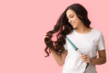 Beautiful young woman with curling iron on pink background