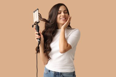 Beautiful young woman with curling iron on beige background