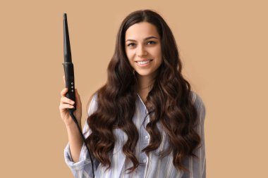 Beautiful young woman with curling iron on beige background
