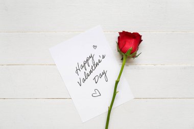 Card with text HAPPY VALENTINE'S DAY and red rose on white wooden background