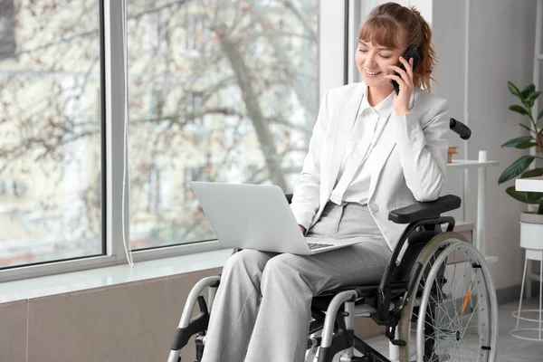 Young businesswoman in wheelchair with laptop talking by mobile phone at office