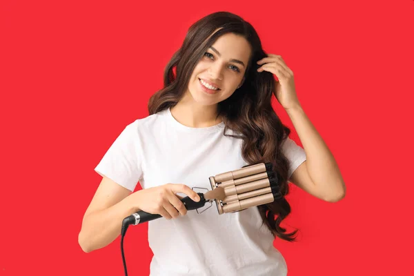 Beautiful young woman with curling iron on red background