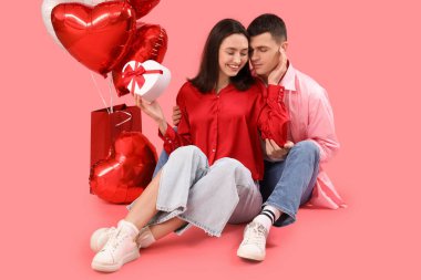 Young couple with gift and heart-shaped balloons sitting on pink background. Valentine's Day celebration