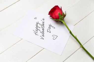Card with text HAPPY VALENTINE'S DAY and red rose on white wooden background