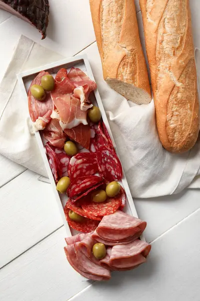 Plate with assortment of tasty deli meats and baguette on white wooden background