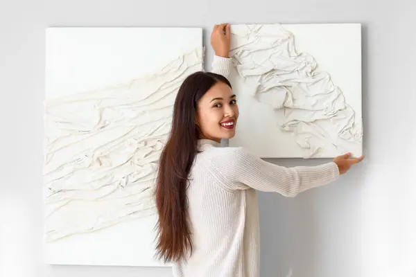 Young Asian woman hanging 3D painting on light wall at home