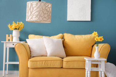 Oturma odasındaki masada minderleri ve çiçek açan narsist çiçekleri olan rahat sarı kanepe.