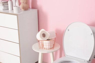 Basket with paper rolls, toilet bowl and chest of drawers near color wall, closeup