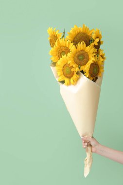Female hand with bouquet of beautiful sunflowers in paper on green background