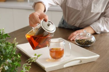 Genç bir kadın mutfakta cam bardağa sıcak çay dolduruyor.