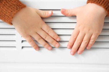 Evdeki radyatörün yanında elleri ısıtan sevimli küçük kız, yakın plan.