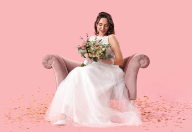 Beautiful bride with wedding bouquet in armchair on pink background