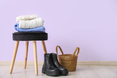 Stacked clothes on stool with basket and boots near lilac wall