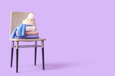 Stacked sweaters with hat on chair against lilac background