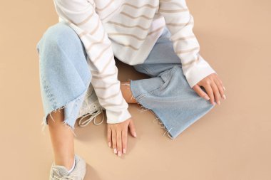 Young woman in trendy jeans sitting on beige background, closeup