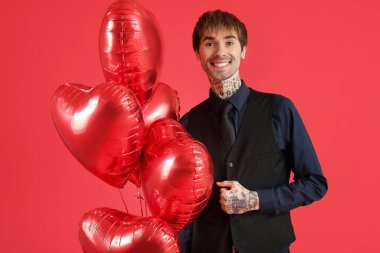 Handsome young man with heart-shaped air balloons for Valentine's day on red background