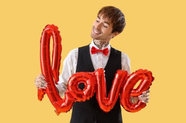 Handsome young man with air balloon in shape of word LOVE on yellow background. Valentine's day celebration