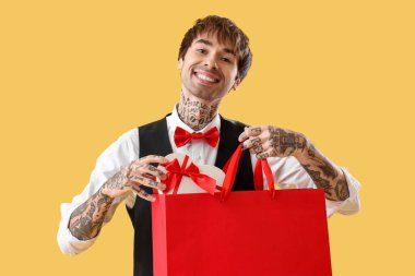 Handsome young man putting gift for Valentine's day in shopping bag on yellow background