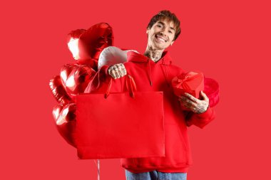 Handsome young man with heart-shaped air balloons, shopping bag and gifts for Valentine's day on red background